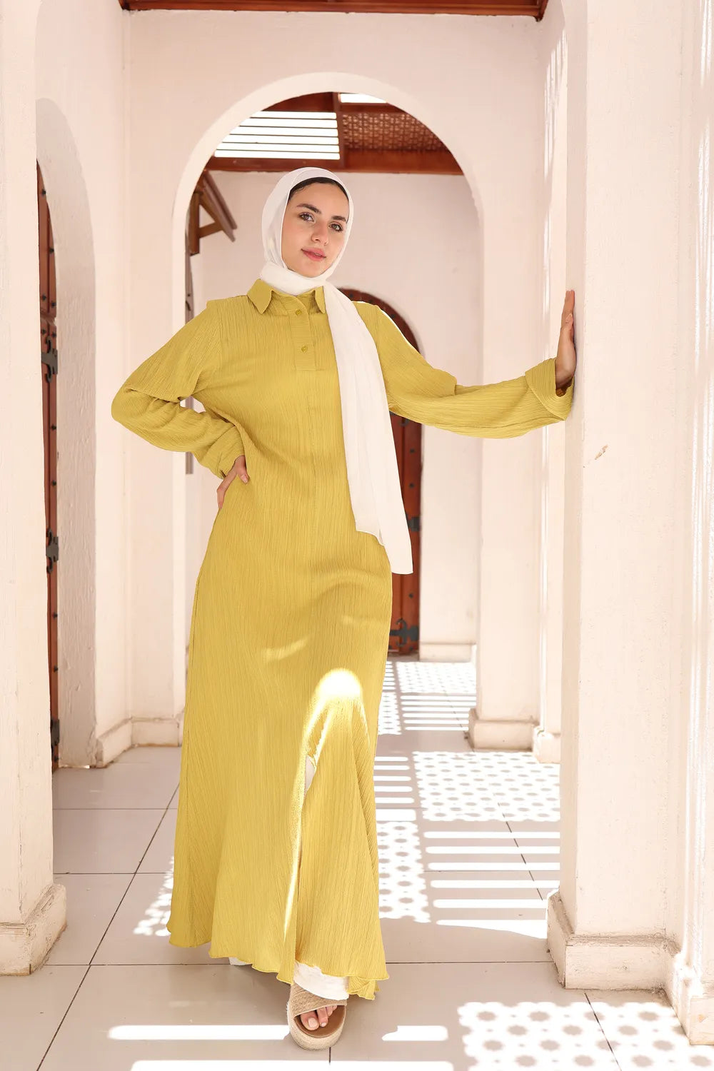Yellow modest long dress worn by hijabi model posing with one hand on the wall
