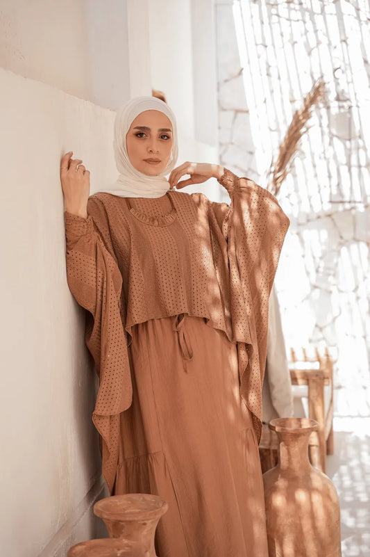 Woman in a brown tunic poncho with modest dress standing against a white wall with decorative elements.