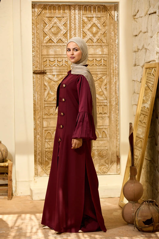 A woman wearing a long burgundy Longline Sleeveless Zip Vest with gold buttons and a light beige hijab stands in front of an ornate carved wooden door in a warm, rustic interior.