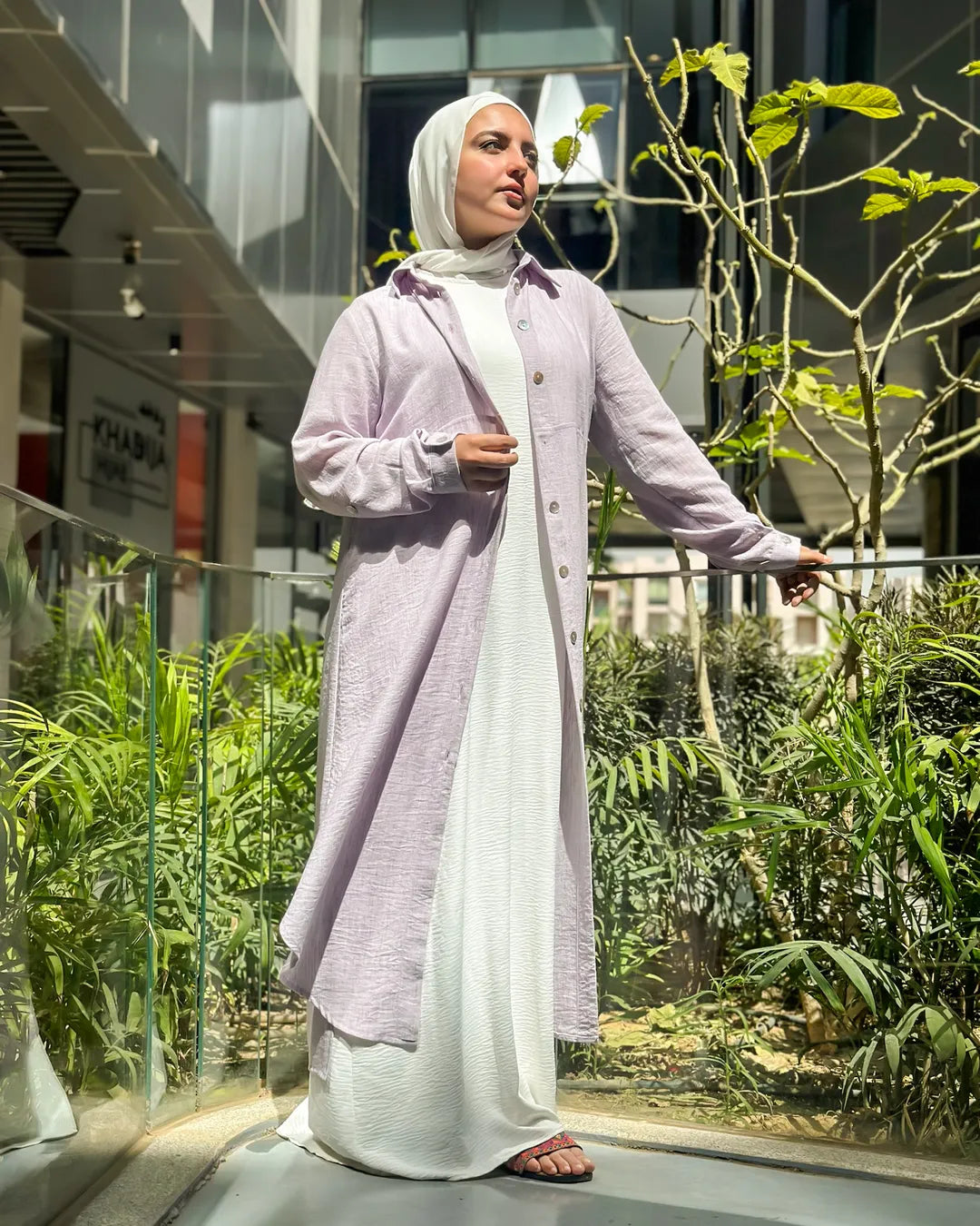 Hijabi model wearing a soft lilac full-length modest dress, posing in an urban outdoor walkway