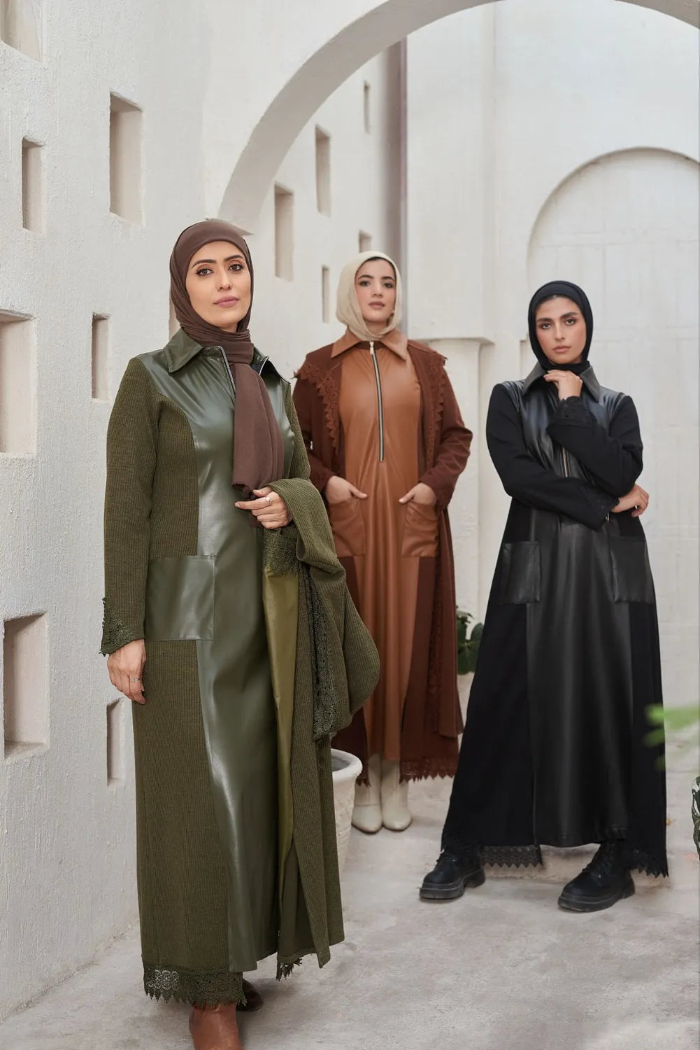 Three women in hijabs  and leather dresses standing in a row against a white architectural background