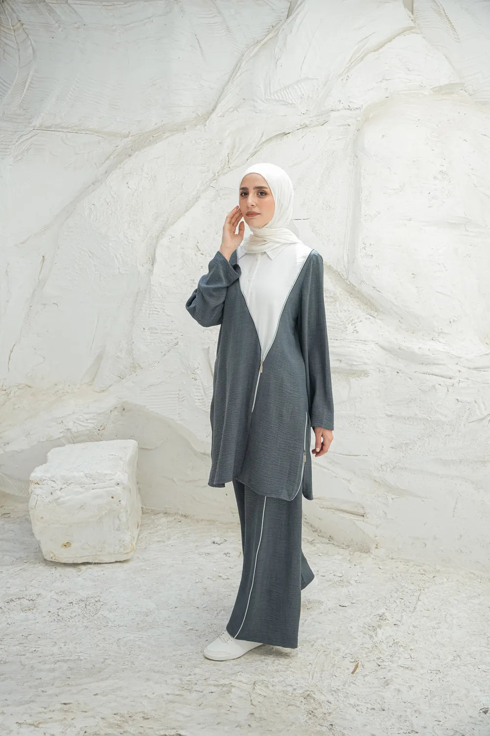 Woman wearing a gray outfit with a white hijab against a textured white wall.