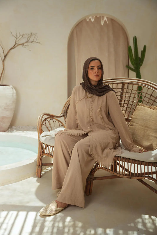 Woman in a beige modest set with long tunic top and wide-leg pants, seated on a rattan chair in a sunlit courtyard by the pool