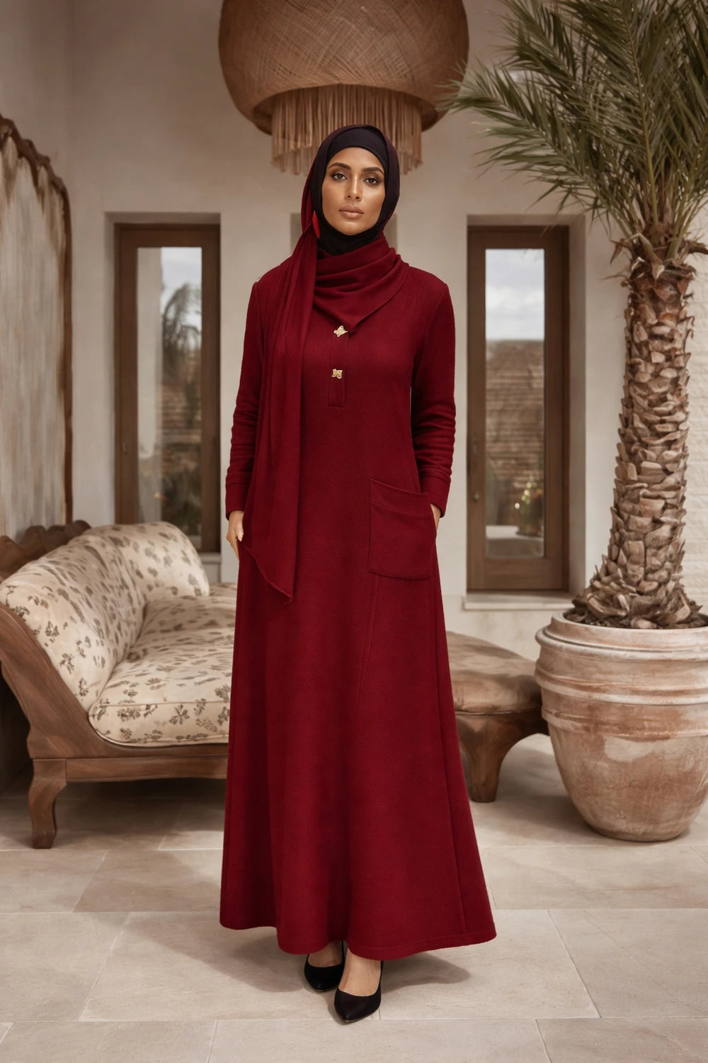 Woman  in a hijab in a redlong  dress standing in a room with a couch and palm tree.