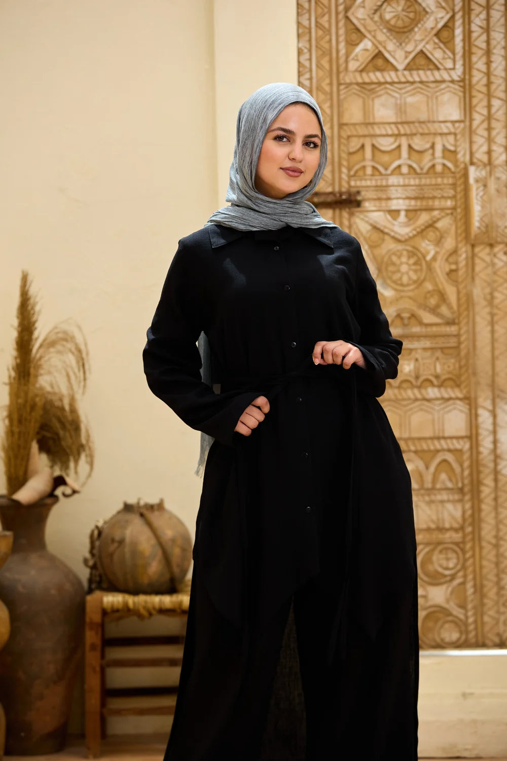 A woman wearing a black long-sleeve tunick and a light gray hijab, standing in front of a beige wall with a carved wooden door and decorative vases.