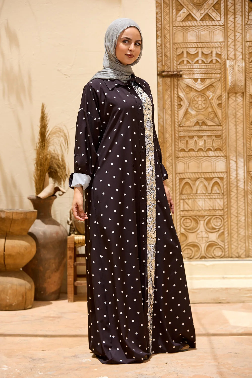 A woman wearing a long black abaya with white polka dots and a silver embroidered front panel, paired with a light gray hijab, standing in front of a carved wooden door.
