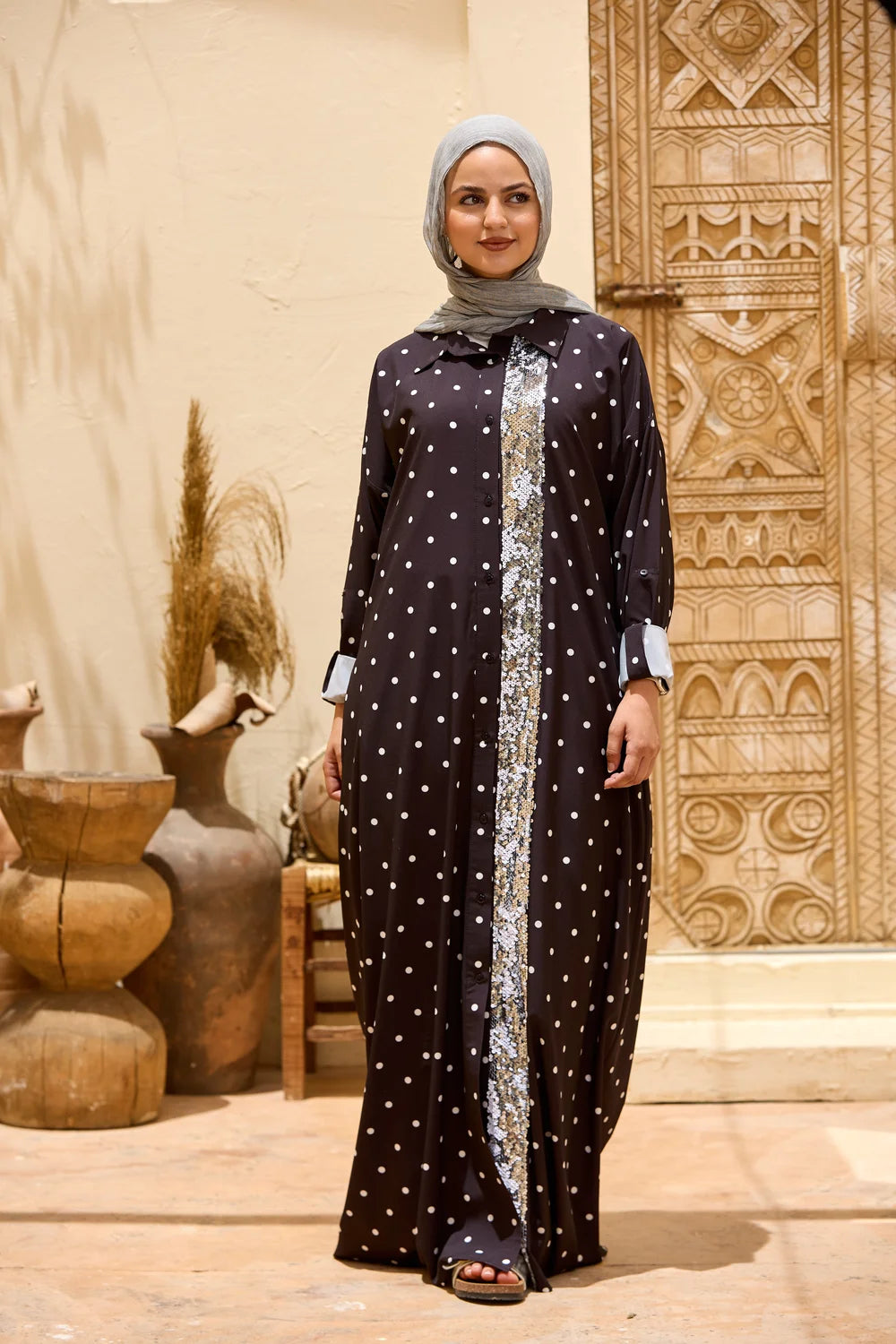 A woman wearing a long black modest dress with white polka dots and with a sequin front panel, paired with a gray hijab, standing in front of a carved wooden door and decorative clay pots.