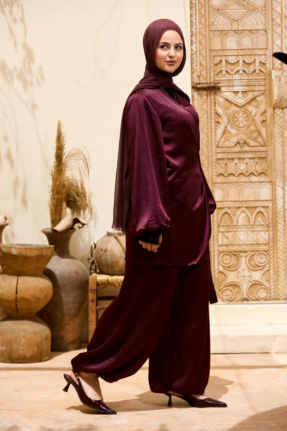 A woman wearing a deep burgundy two-piece modest outfit with a matching hijab, walking in a styled indoor setting with decorative pottery and carved walls.