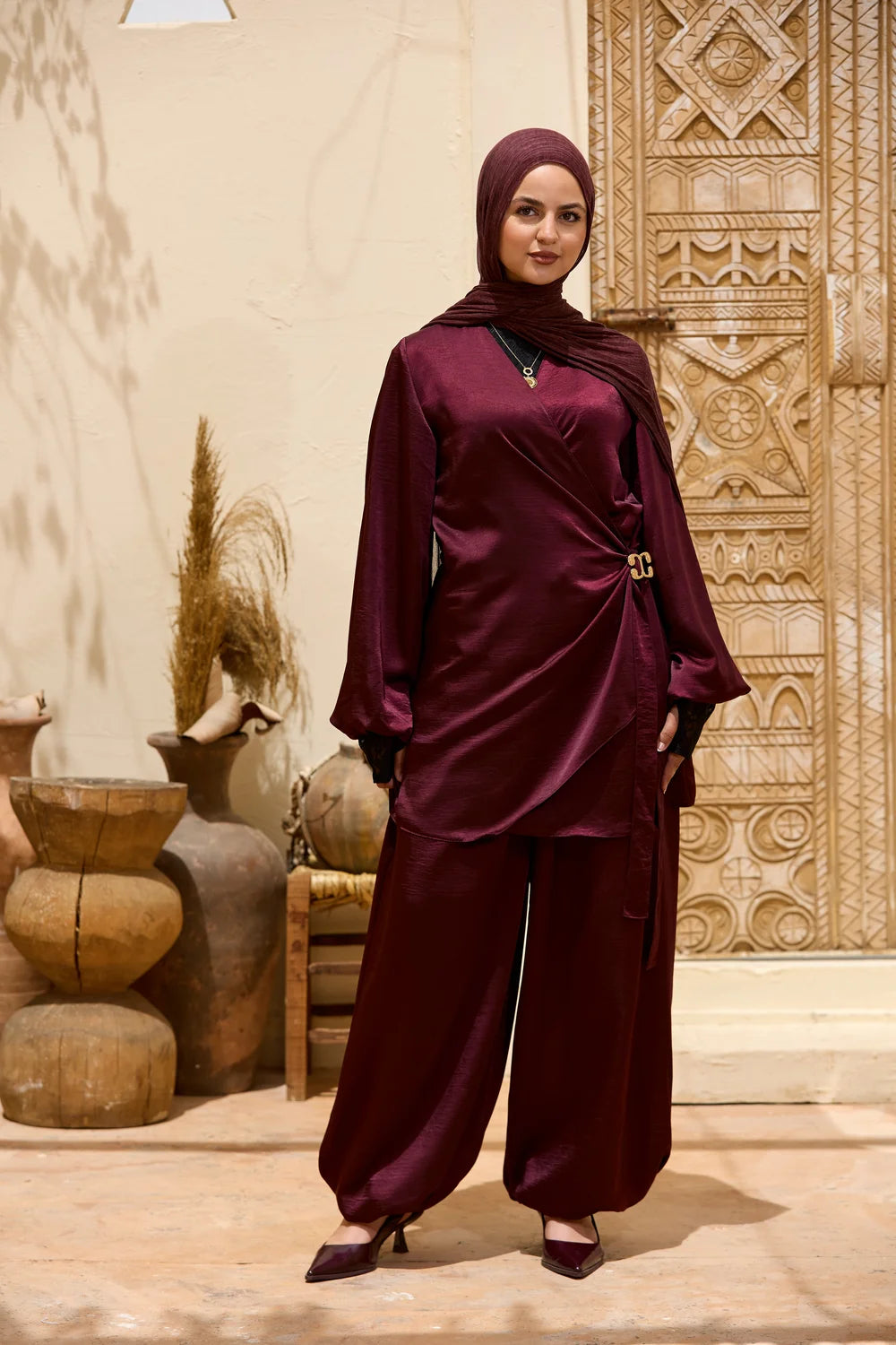 A woman modeling a  burgundy Satin Wrap Suit with Adjustable Balloon Wide-Leg Pantst with a matching hijab, standing in front of a carved wooden door and decorative clay pots.