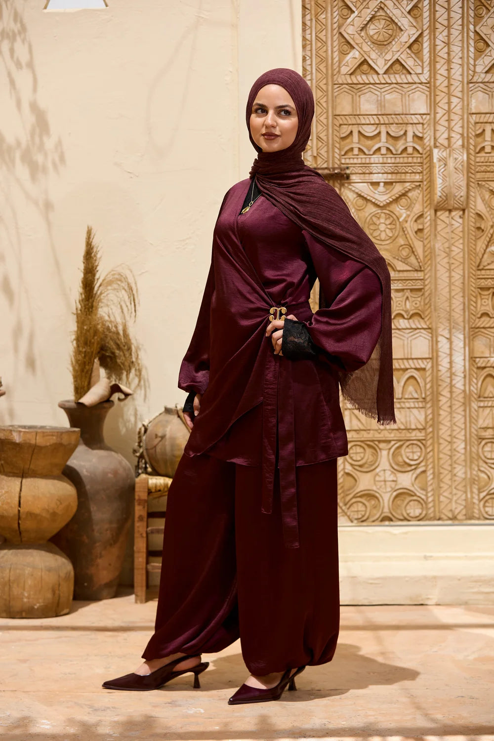 A woman modeling a  burgundy Satin Wrap Suit with Adjustable Balloon Wide-Leg Pants with burgundy hijab, standing in front of a carved beige wall and decorative pottery.