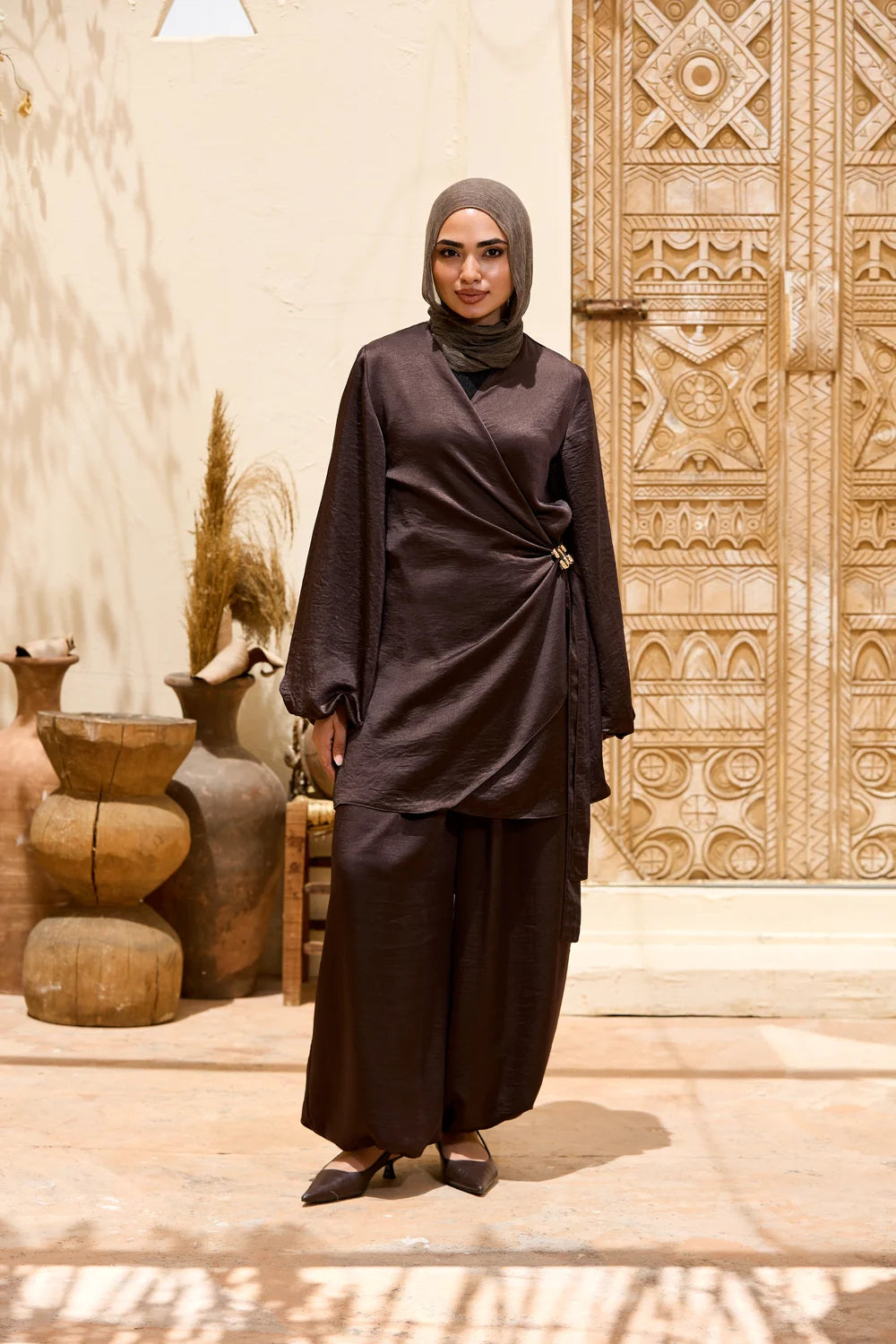 A woman wearing a dark brown hijab and a matching dark brown Satin Wrap Suit with Adjustable Balloon Wide-Leg Pants, standing in front of a carved beige door and rustic pottery.