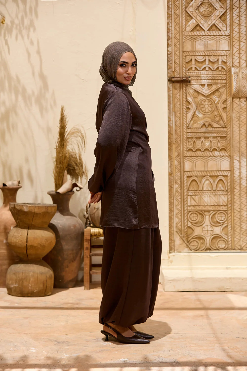 A woman wearing a dark brown modest Satin Wrap Suit with Adjustable Balloon Wide-Leg Pants with a matching hijab stands in profile in front of a carved wooden door and rustic clay pots.