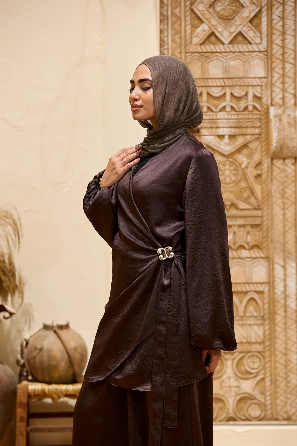A woman wearing a dark brown modest outfit  Set with a matching hijab, standing in front of a beige textured wall with carved patterns.