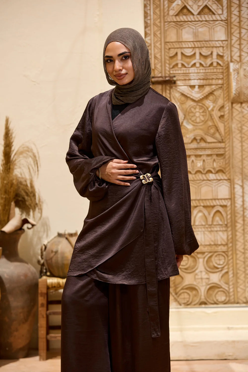 A woman wearing a dark brown, Satin Wrap Suit with Adjustable Balloon Wide-Leg Pants with a matching hijab and a thin metallic belt, standing in front of an ornate beige carved door.