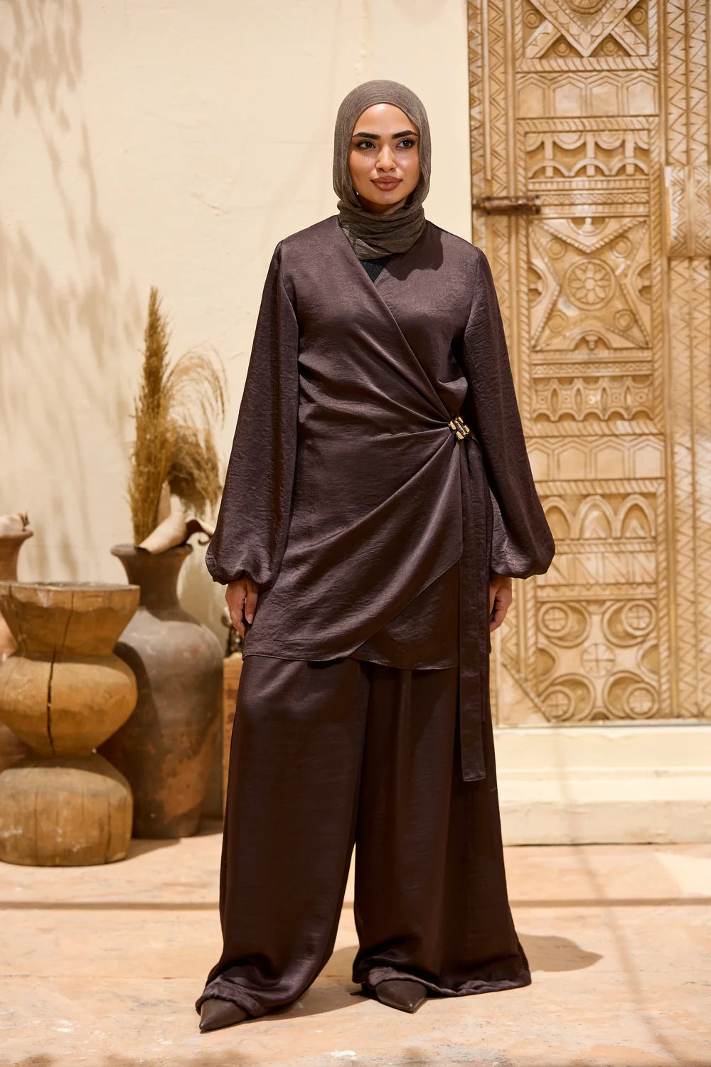 A woman wearing a dark brown two-piece modest outfit  Satin Wrap Suit with Adjustable Balloon Wide-Leg Pantswith a matching hijab, standing in front of a carved beige wall and decorative pottery.