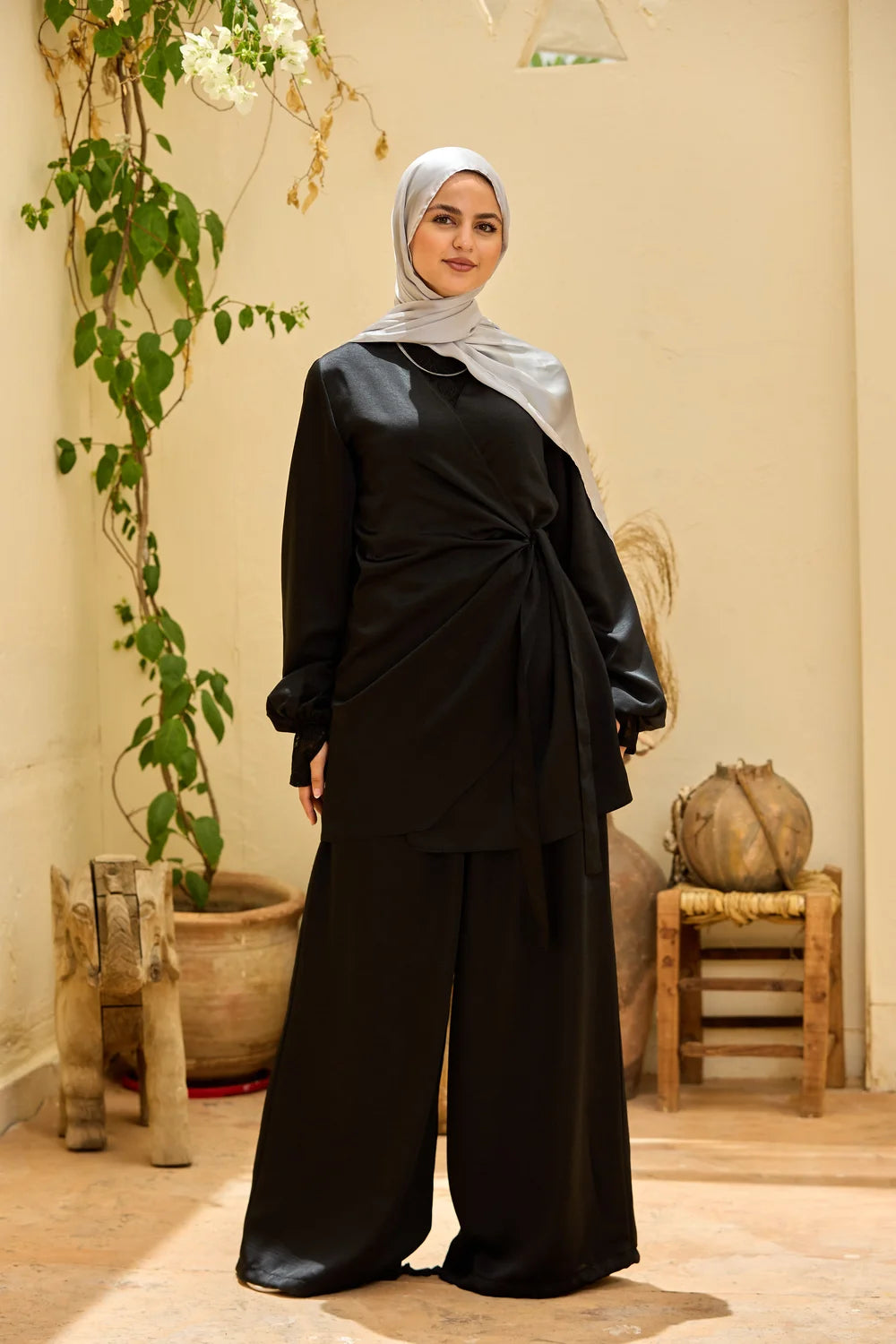 A woman wearing a black Satin Wrap Suit with Adjustable Balloon Wide-Leg Pants, paired with a light gray hijab, standing in a warm-toned indoor setting with plants and rustic decor.
