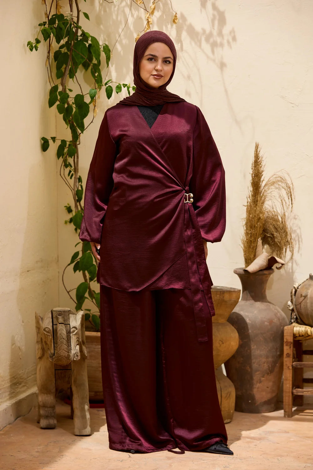 A woman wearing a burgundySatin Wrap Suit with Adjustable Balloon Wide-Leg Pants with a matching hijab, standing in a rustic indoor setting with decorative clay pots and a plant.