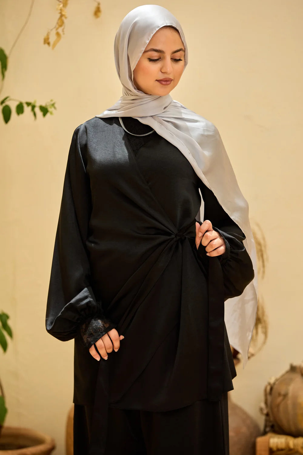 A woman wearing a black Satin Wrap Suit with Adjustable Balloon Wide-Leg Pants and a light gray hijab, standing against a warm beige wall.