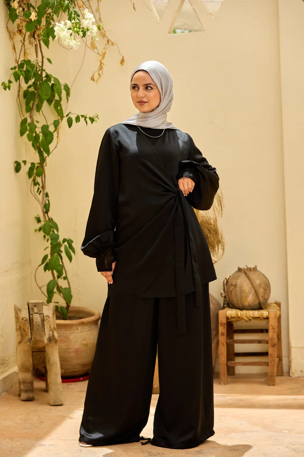 A woman wearing a black satin-Satin Wrap Suit with Adjustable Balloon Wide-Leg Pants, paired with a light gray hijab, standing in a warm-toned indoor setting with plants and wooden furniture.