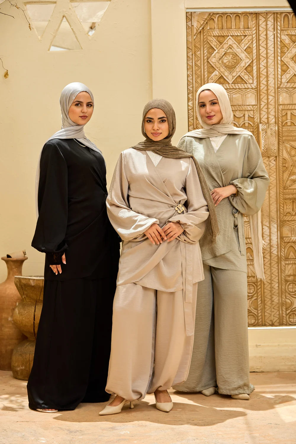Three women wearing modest Islamic clothing Satin Wrap Suit with Adjustable Balloon Wide-Leg Pantsand and  hijabs, standing together in front of a decorative beige wall and carved wooden door.