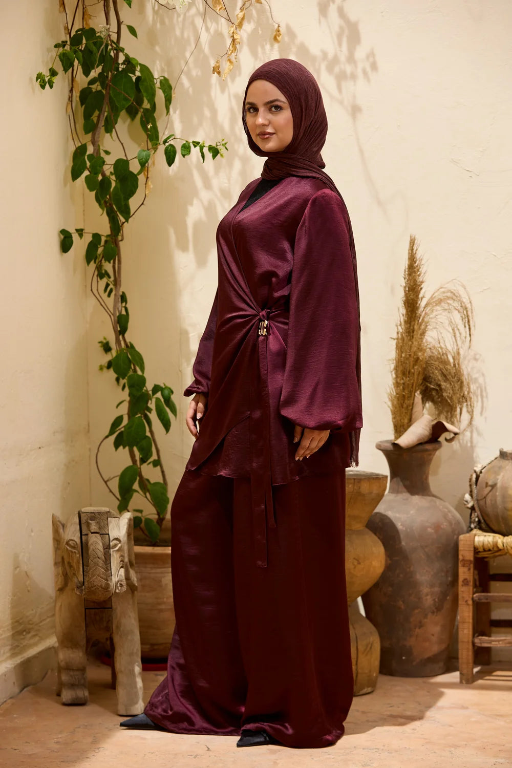 A woman wearing a  burgundy Satin Wrap Suit with Adjustable Balloon Wide-Leg Pants and matching hijab stands in a styled indoor setting with plants and ceramic vases.