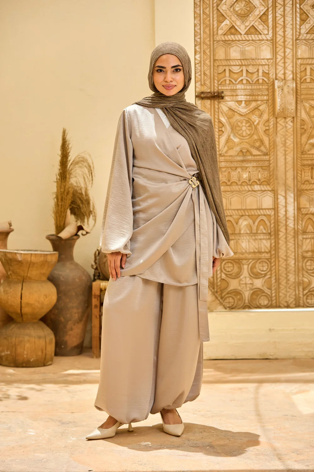 A woman wearing abeige Satin Wrap Suit with Adjustable Balloon Wide-Leg Pants with a matching hijab, standing in front of a carved wooden door and decorative clay pots.