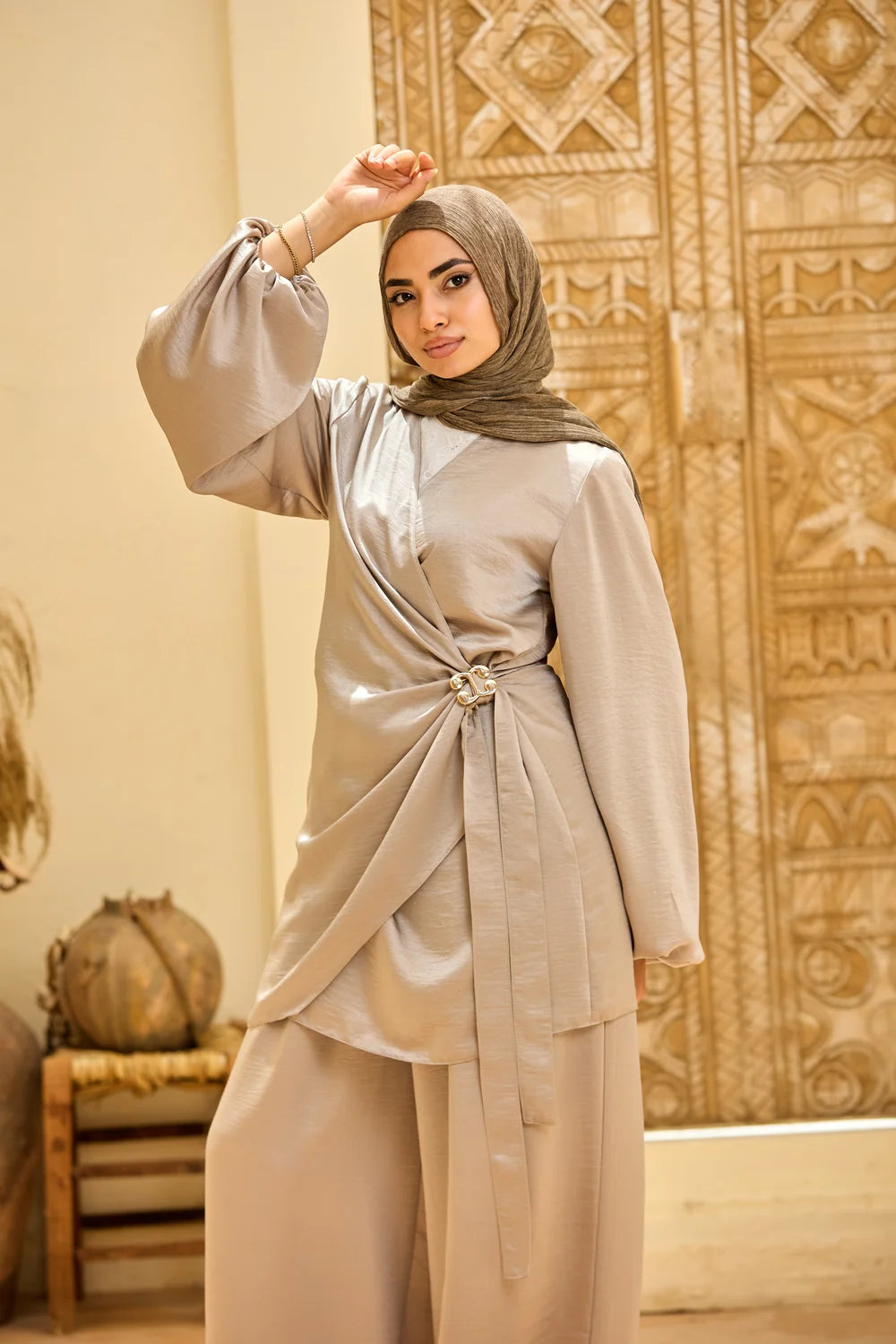A woman wearing a beige lSatin Wrap Suit with Adjustable Balloon Wide-Leg Pants with a matching hijab, standing in front of a patterned beige wall.