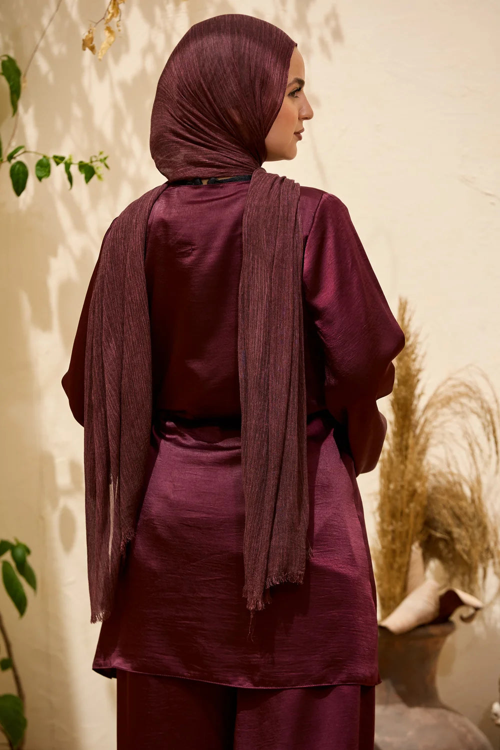 A woman wearing a burgundy Satin Wrap Suit with Adjustable Balloon Wide-Leg Pants with a matching hijab, shown from the back in a softly lit indoor setting with decorative plants and dried grass.