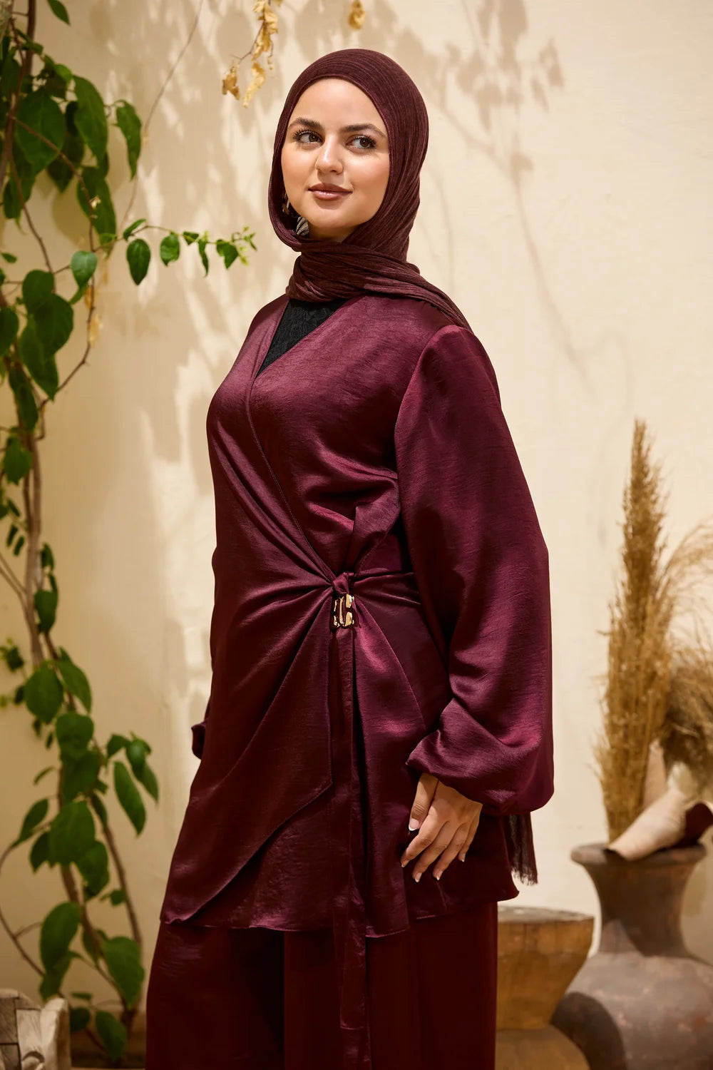 A woman wearing a  burgundy Satin Wrap Suit with Adjustable Balloon Wide-Leg Pants with a matching hijab, standing in a styled indoor setting with plants and dried grass.