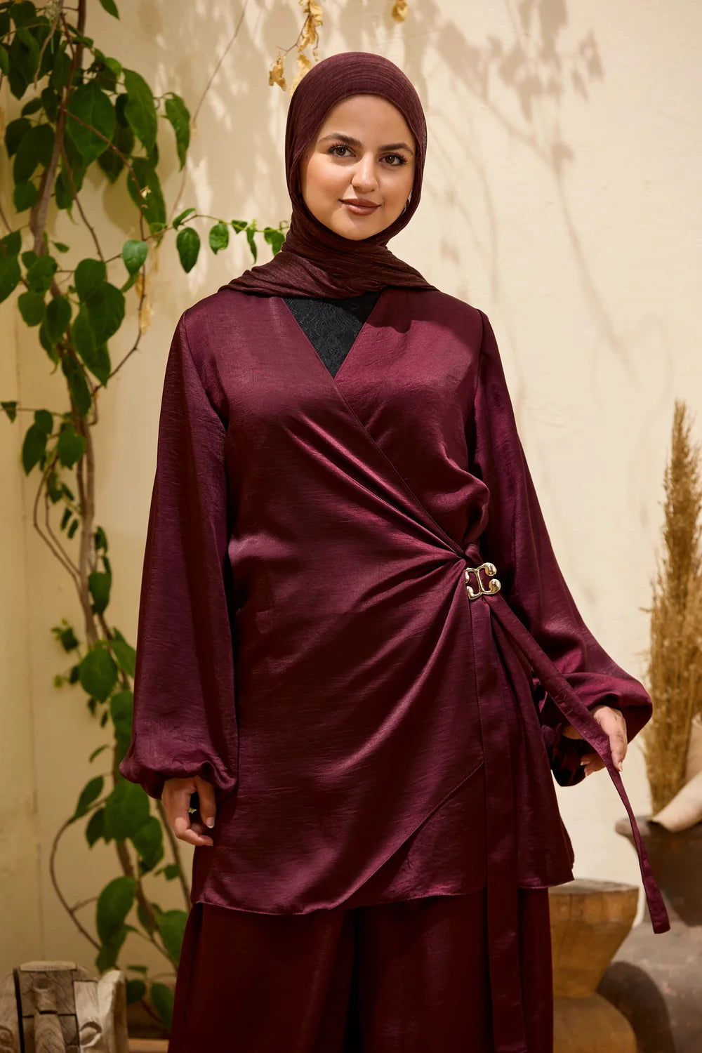 A woman wearing a  burgundy satin hijab and Satin Wrap Suit with Adjustable Balloon Wide-Leg Pants, styled with a black inner top. She is standing in an indoor setting with a beige wall, green climbing vines, and dried plants.