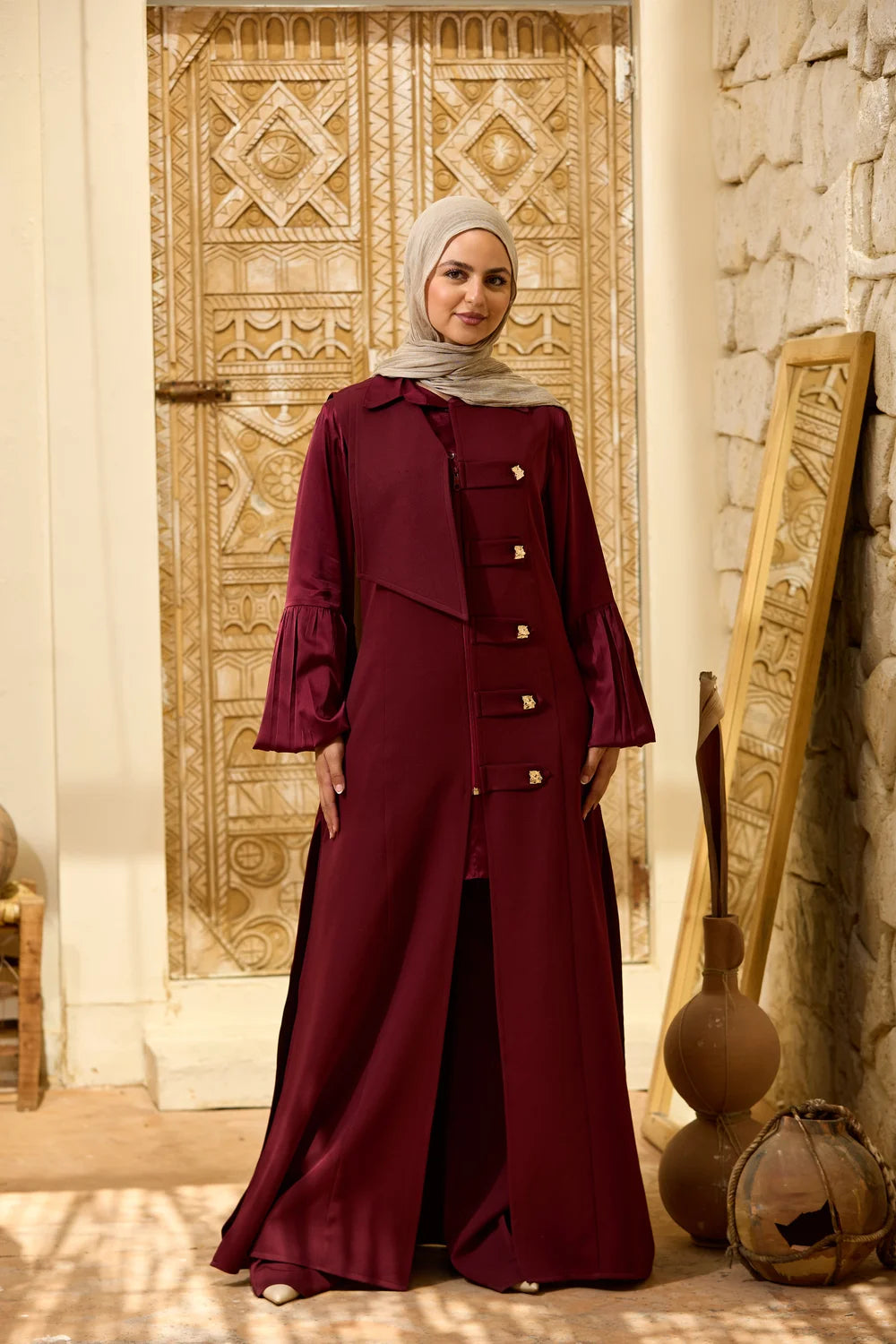A woman wearing a long burgundy Longline Sleeveless Zip Vest with gold-tone buttons and a light gray hijab, standing in front of an ornate carved wooden door in a warm, rustic interior.