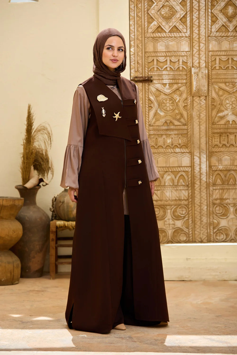 A woman wearing a long dark brown Longline Sleeveless Zip Vest with a matching hijab, standing in front of an ornate carved wooden door. The Longline Sleeveless Zip Vest has a structured vest-style front with decorative gold-toned embellishments and buttons, worn over a sheer taupe long-sleeve underlayer.