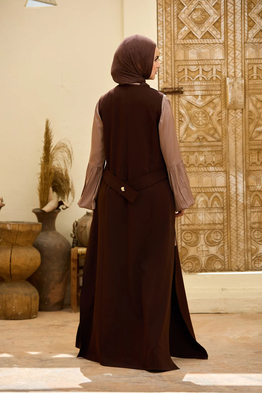 A woman wearing a  dark brown Longline Sleeveless Zip Vest  with  a matching brown hijab, photographed from behind in an indoor setting with a carved wooden door and decorative vases.