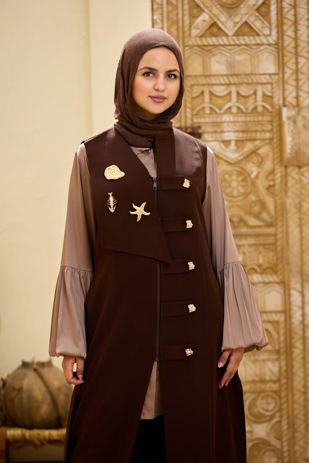 A woman wearing a dark brown Longline Sleeveless Zip Vest with gold decorative accents and a matching brown hijab, standing in front of an ornate beige and gold patterned wall.
