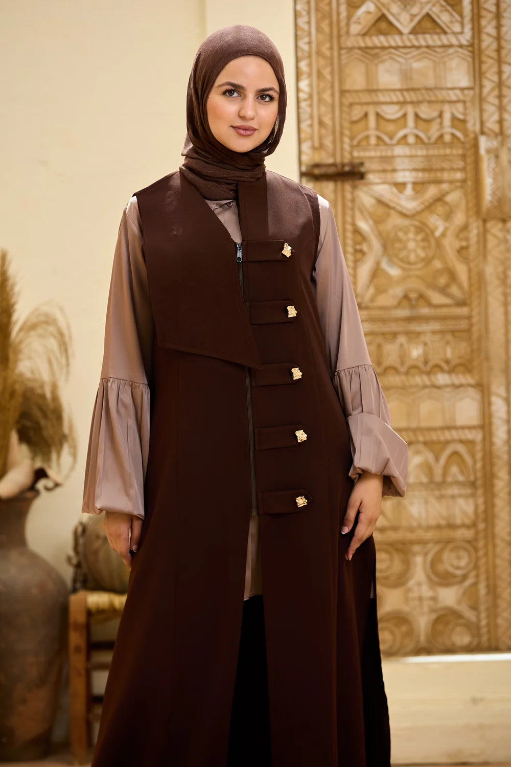 A woman wearing a  dark brown Longline Sleeveless Zip Vest dress with decorative gold-tone buttons, a matching brown hijab, and a light taupe long-sleeve blouse, standing in front of an ornate carved wooden door.