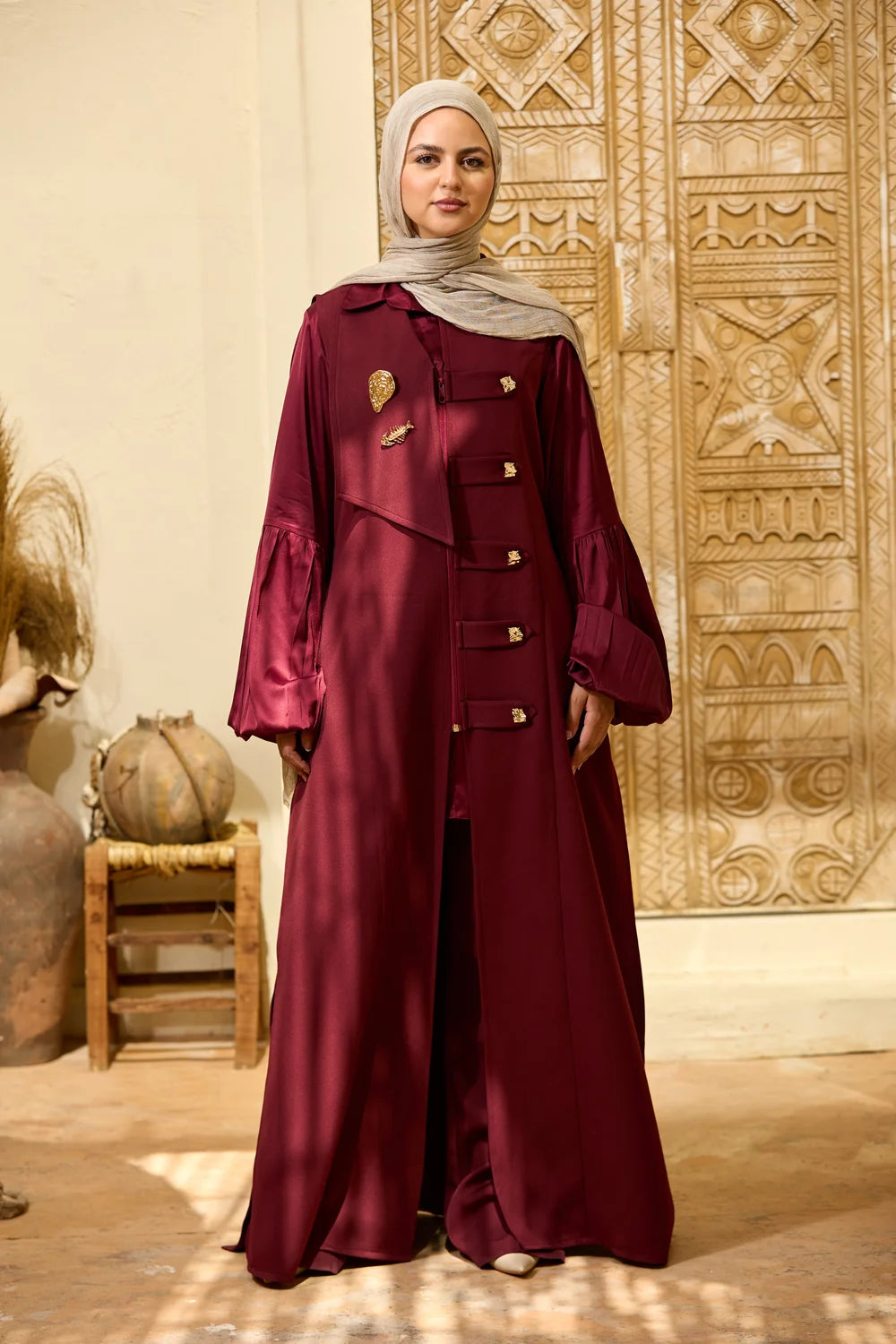 A woman wearing a long, игкпгтвн Longline Sleeveless Zip Vest with gold-tone buttons and a light gray hijab, standing in front of an ornate carved wooden door.
