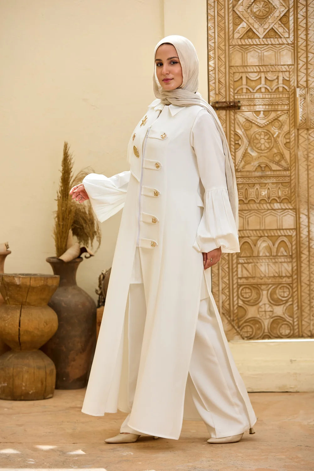 A woman wearing a Longline Sleeveless Zip Vest with gold buttons, matching wide-leg pants, and a white hijab, standing in front of a beige wall and an ornate carved wooden door.