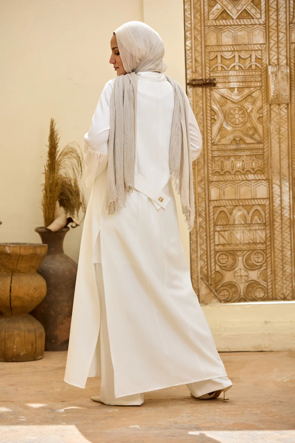 A woman wearing aLongline Sleeveless Zip Vest and matching wide-leg trousers, paired with a light beige hijab, walking in front of a carved wooden door.