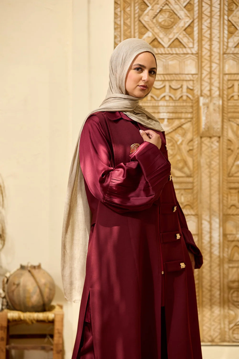 A woman wearing a burgundy Longline Sleeveless Zip Vest  with gold button accents and a light beige hijab, standing in front of an ornate carved wooden door.