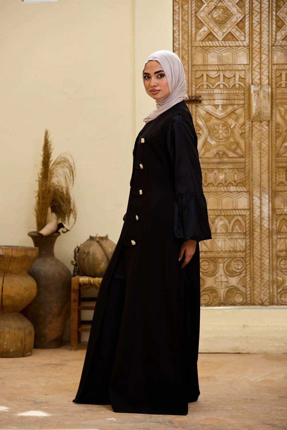 A woman wearing a black Longline Sleeveless Zip Vest with gold buttons and a light gray hijab, standing in a decorated interior with a carved wooden door and clay vases.