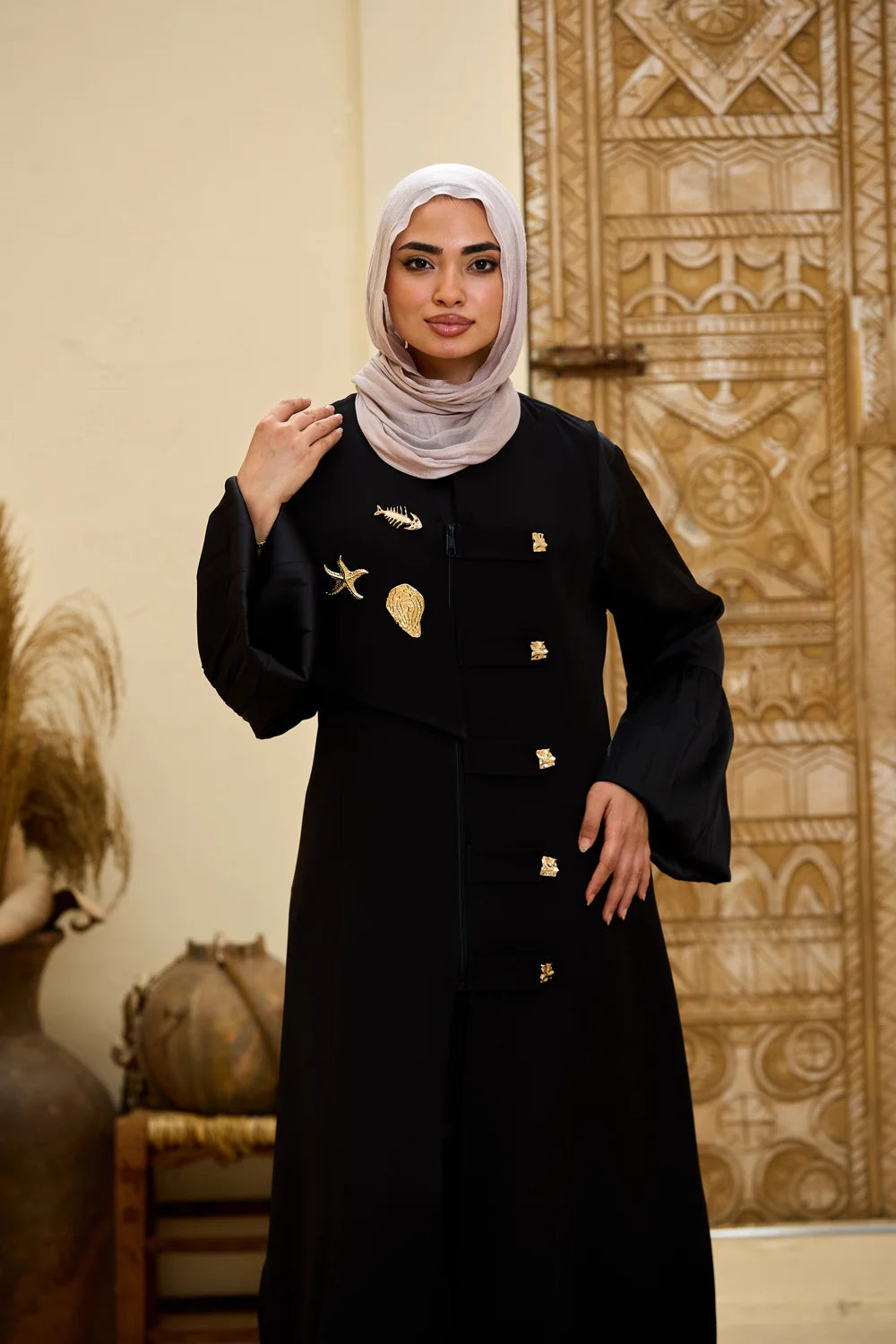 A woman wearing a black Longline Sleeveless Zip Vest with gold decorative details and a light beige hijab, standing in front of a carved wooden door.