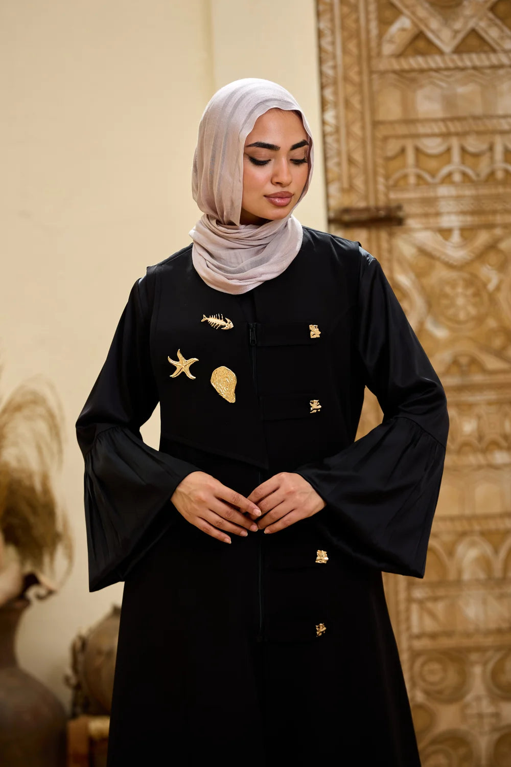 A woman wearing a black Longline Sleeveless Zip Vest with gold decorative details and a light beige hijab, standing in front of an ornate beige background.