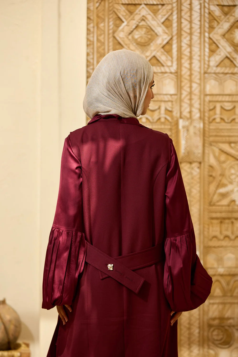 A woman viewed from behind wearing a Longline Sleeveless Zip Vest with a matching waist tie and a light beige hijab, standing in front of an ornate carved wooden door.