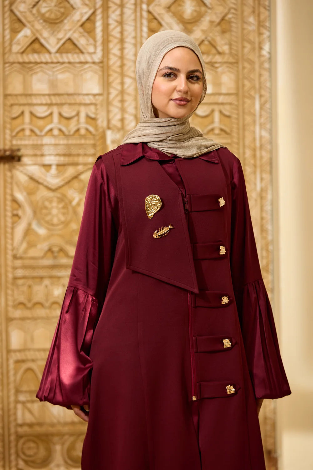 A woman wearing a burgundy Longline Sleeveless Zip Vest with gold decorative buttons and brooch details, paired with a light beige hijab, standing in front of an ornate gold-toned patterned door.