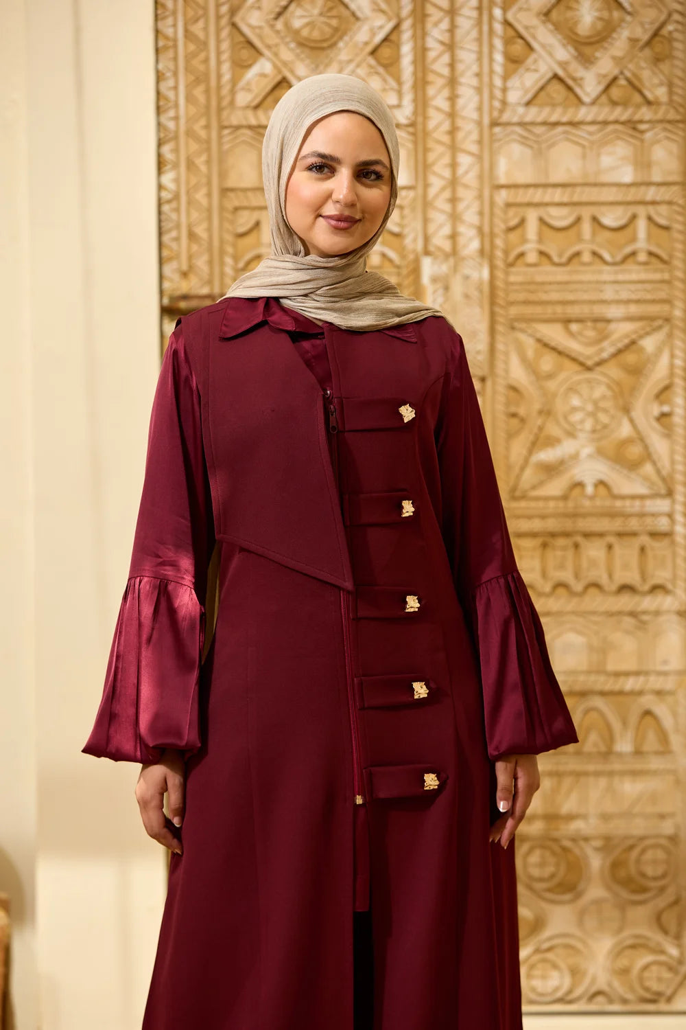 A woman wearing a  burgundyLongline Sleeveless Zip Vest with gold-tone decorative buttons and a light beige hijab, standing in front of an ornate beige patterned wall.