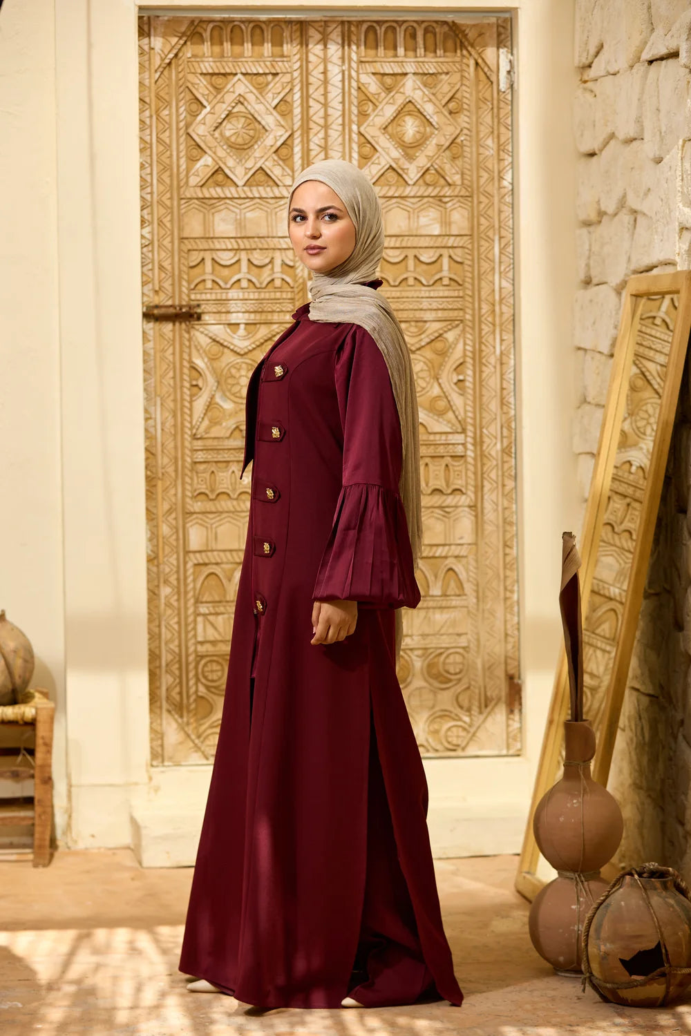 A woman wearing a long burgundy Longline Sleeveless Zip Vest with gold buttons and a light beige hijab stands in front of an ornate carved wooden door in a warm, rustic interior.