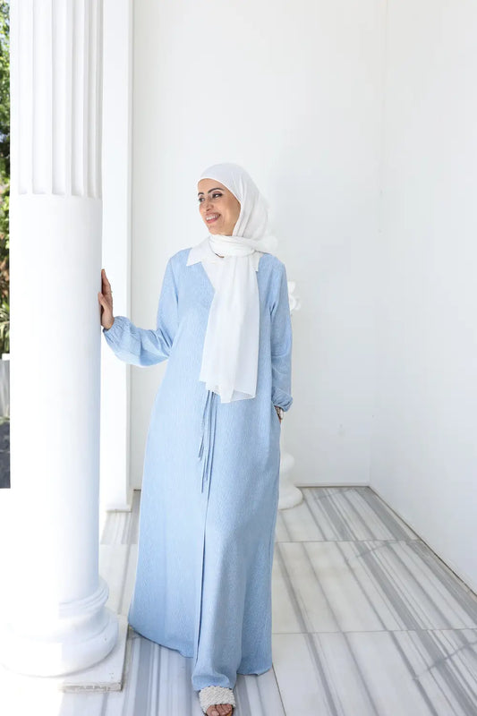 Woman wearing a light blue dress  and white hijab standing against a white wall.
