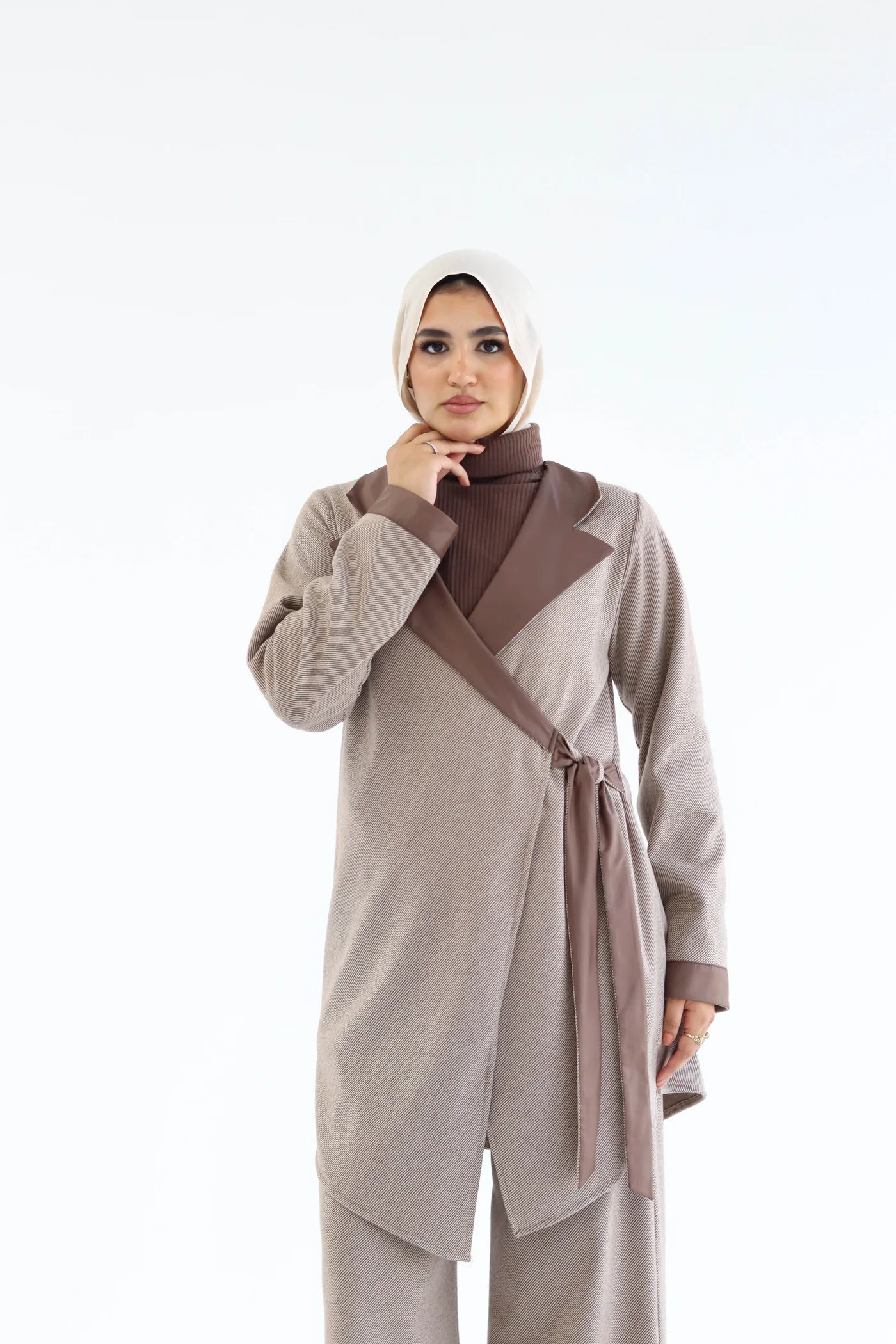 Woman wearing a beige outfit with a brown sash on a white background
