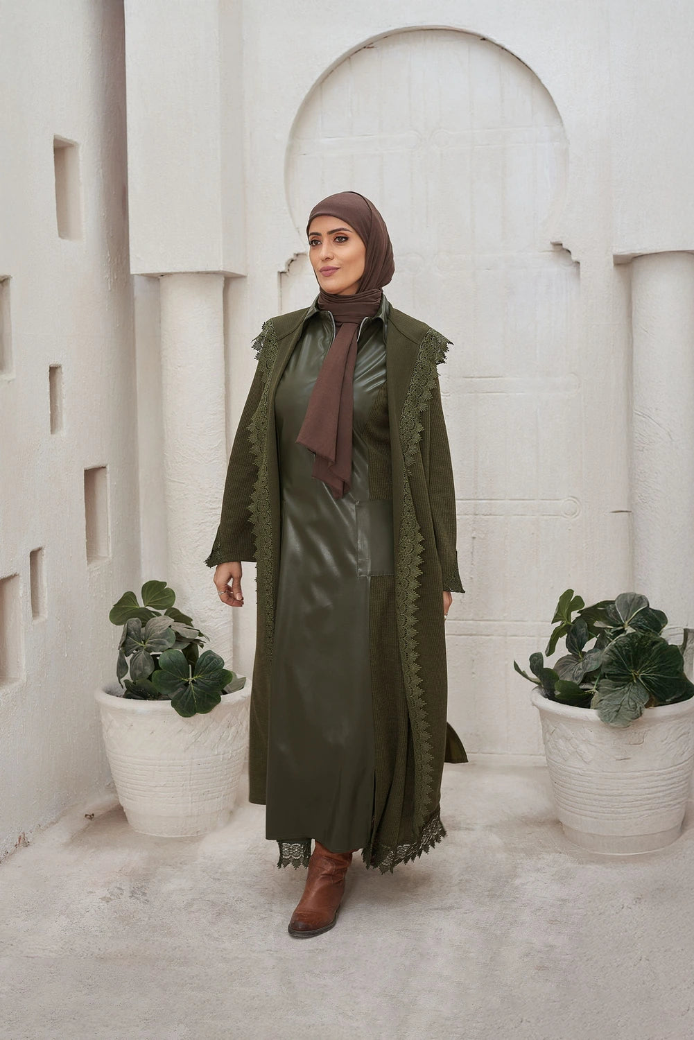 Woman in a green dress and Long knit guipure cardigan with leather details for modest winter layering standing in front of a white architectural background with plants.