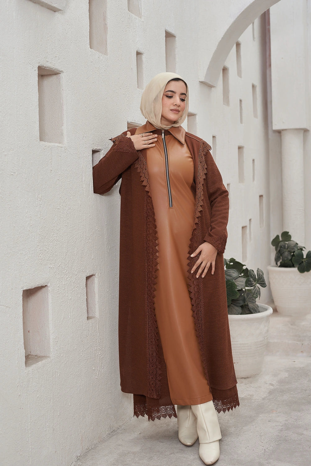 Woman in a brown Long knit guipure cardigan with leather details for modest winter layering  standing against a light-colored wall with architectural elements.